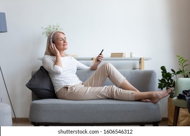 Calm Mature Woman Relax On Cozy Couch At Home Wear Earphones Enjoy Music Using Modern Cellphone, Modern Senior Female Gadget User In Headphones Listen To Favorite Track Good Sound On Smartphone