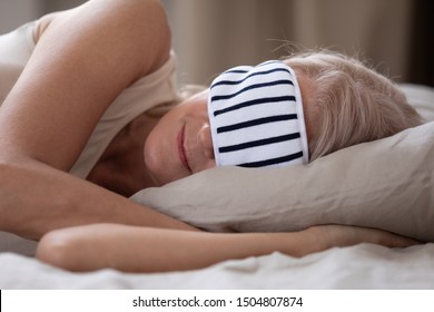 Calm Mature Woman Wearing Striped Sleeping Mask Relaxing In Comfortable Bed Close Up, Peaceful Beautiful Older Female Resting In Bedroom, Enjoying Fresh Bedclothes, Lying On Soft Pillow