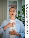 Calm middle aged woman practicing breathing meditation techniques standing with closed eyes, self caring at home. Focused on mental health female holding hands on chest and belly doing pranayama yoga.