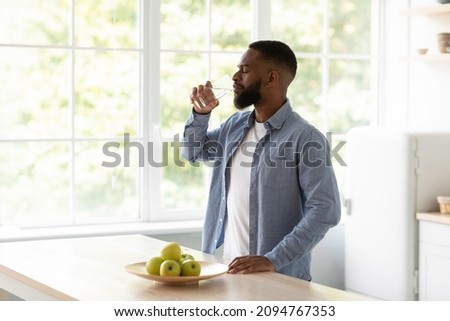 Calm millennial african american bearded man with closed eyes drinks water from glass in kitchen interior with apples on window background. Thirst, aqua balance, healthy nutrition and hydration