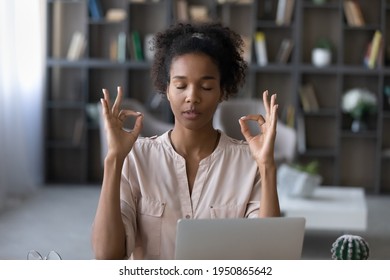 Calm millennial African American woman distracted form computer work meditate at home office desk. Young biracial female relax practice yoga at workplace, breathe fresh air. Peace concept.