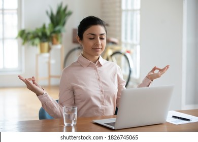 Calm millennial indian female employee sit at office desk distracted from computer work meditating. Young ethnic woman practice yoga at workplace, relieve negative emotions. Stress free concept. - Powered by Shutterstock