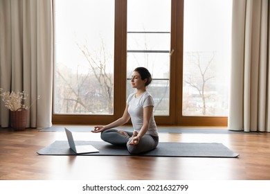 Calm millennial Indian woman sit on mat at home meditate watch online training or session on laptop. Peaceful young ethnic female practice yoga breathe fresh air have webcam call with trainer.