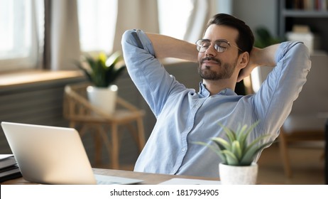 Calm millennial man in glasses sit relax at home office workplace take nap or daydream. Happy relaxed Caucasian young male rest in chair distracted from computer work, relieve negative emotions.