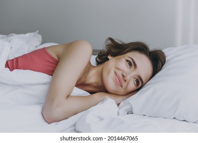 Calm minded caucasian happy young woman 20s wear pajamas tank shirt lying in bed lies wrap covered under blanket duvet on pillow rest relax indoors at home. Good mood night morning bedtime concept.