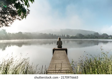 Calm misty morning relaxation by pond.Sitting woman in countryside.Intended female mysterious atmosphere. Spring foggy nature.Silence.Woman feeling freedom,enjoying vacation.No stress,calm mind,relax