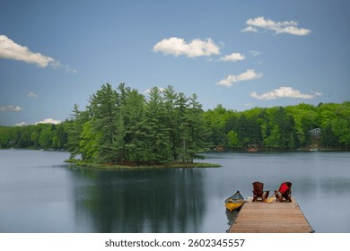 The calm Muskoka lake reflects the sky as it stretches toward a tree-covered island. A wooden dock with two Adirondack chairs, a yellow canoe, and an oar rests peacefully along the shore. - Powered by Shutterstock - Get 15% off with code: PIKWIZARD15