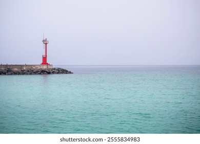 A calm ocean scene with a vibrant red lighthouse standing on a rocky pier against soft pastel skies. The turquoise water and minimalistic composition highlight tranquility and coastal beauty - Powered by Shutterstock