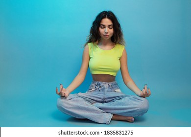 Calm Peaceful Beautiful Girl Sits Resting In Easy Pose, Her Hands In Mudra. Girl Meditating Isolated Over Bright Blue Background. Practicing Yoga Concept. Mental Wellbeing Concept