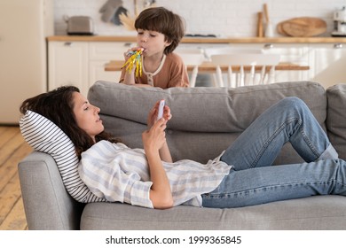 Calm Peaceful Mom Or Nanny Lying On Sofa With Mobile Phone Distracted From Noisy Loud Son Drawing Attention. Young Mother Relax At Home With Smartphone Rest After Work Or Parent Routine On Lockdown