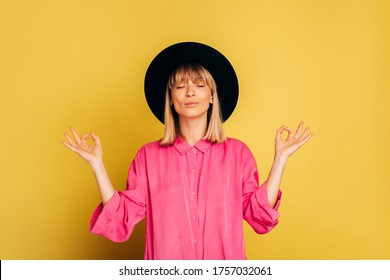 Calm Peaceful Stylish Young Woman Meditating With Closed Eyes. Trendy Girl With Short Blonde Haircut Posing On Camera. Fashionable Model In Pink Shirt And Black Hat Isolated Over Yellow Background