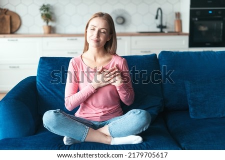 Calm peaceful woman in casual clothes hold hands at chest close to heart, smiling grateful. Female feel thankful, show appreciation and love, sitting on sofa in cozy living room