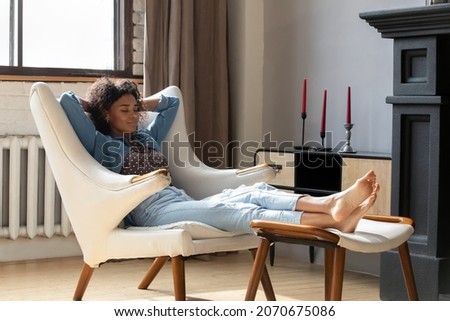 Calm peaceful young African woman taking deep breath of fresh air, reloading mind, resting with closed eyes in soft white armchair. Girl relaxing at home, practicing stress relief techniques