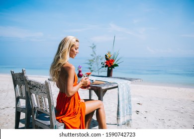  Calm Pensive Adult Blond Lady In Orange Dress Looking Down While Sitting Alone At Wooden Table With Flowers And Tablecloth On Old Chair And Holding Glass Of Red Beverage On Ocean Shore At Bali Resort