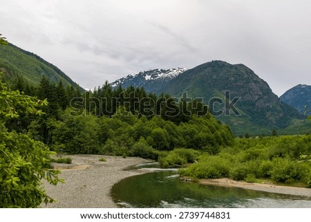 The calm Ralph River flows toward Buttle Creek beside a rocky beach, with forested mountains in the distance under a cloudy sky.