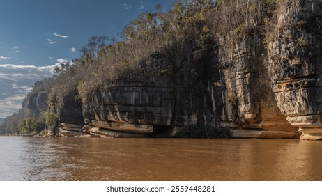 A calm red-brown African river. Trees grow on the steep limestone banks. Blue sky, clouds. Madagascar. Manambolo river. - Powered by Shutterstock