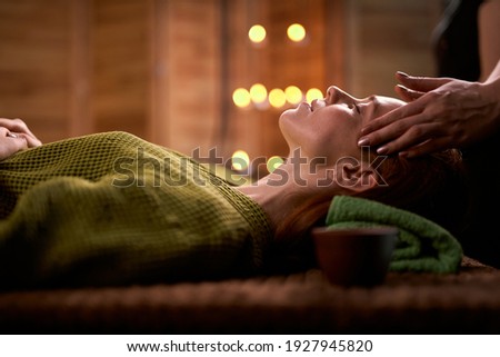 calm redhead woman getting spa treatment isolated in candlelight room, side view on calm female lying on bed in bathrobe enjoying and relaxing