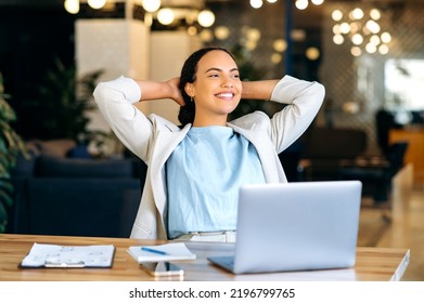 Calm relaxed mixed race female employee resting during work time, sits in modern office, puts hands behind head, feels satisfied by project done, job promotion, looking in distance, smiling friendly