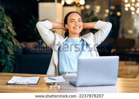 Calm relaxed positive brazilian or hispanic young woman, secretary, manager, financial director, sitting at a desk in a modern office, resting during work time, looking at the camera, smiling friendly