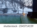 A calm, relaxing winter scene of a chartered open-air hot bath of natural thermal spring, which is lighted up at dusk and located in the forest on a snowy hillside, in an Onsen Ryokan in Iwate, Japan