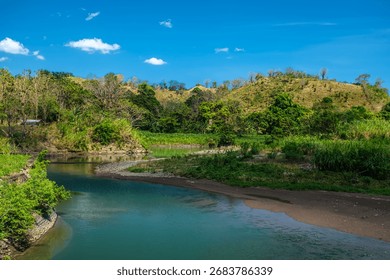 A calm river flows gently between green hills, reflecting the beauty of nature’s balance and serenity in a tropical landscape.