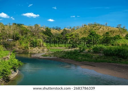 A calm river flows gently between green hills, reflecting the beauty of nature’s balance and serenity in a tropical landscape.