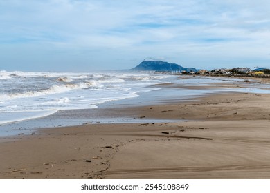 Calm sandy beach after storm; waves roll in, mountain and coastal buildings under partly cloudy sky with scattered driftwood. - Powered by Shutterstock