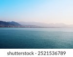 A calm sea in the foreground with a distant view of the mountains partially covered in mist. Clear sky with a light gradient of blue.