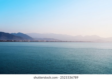 A calm sea in the foreground with a distant view of the mountains partially covered in mist. Clear sky with a light gradient of blue. - Powered by Shutterstock