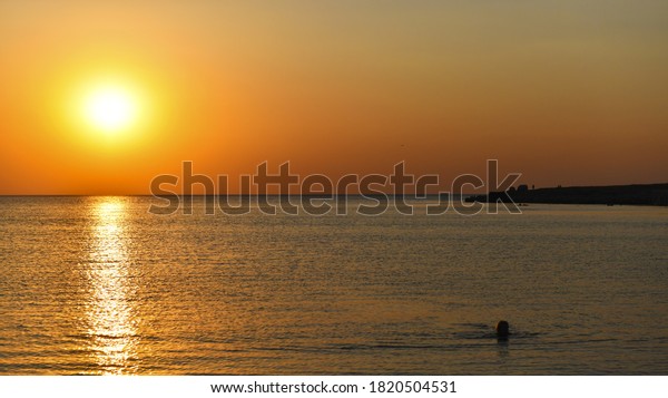 Calm\
sea at sunset. The sun sets over the horizon. People and their car\
on a rocky promontory. The girl swims to the\
shore.