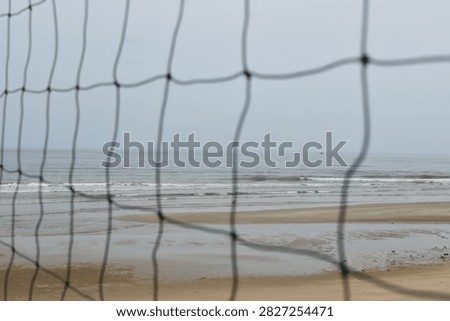A calm seaside beach viewed through a beach volleyball net after rainfall, with gentle waves moving toward the wet sandy shore beneath a dark, overcast sky. The empty volleyball area creates a quiet and melancholic atmosphere, suggesting that outdoor recreation and cheerful beach activities have ended or been interrupted by the rain. The muted gray tones, still ocean, and abandoned recreational setting evoke feelings of loneliness, nostalgia, silence, and the fading excitement of a beach day after bad weather.