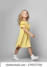 Calm And Self-confident Blonde Curly Kid Girl In Yellow Dress And Sneakers Walks, Passes Us Looking Up Over Grey Background. Stylish Comfortable Everyday Fashion For Children Concept