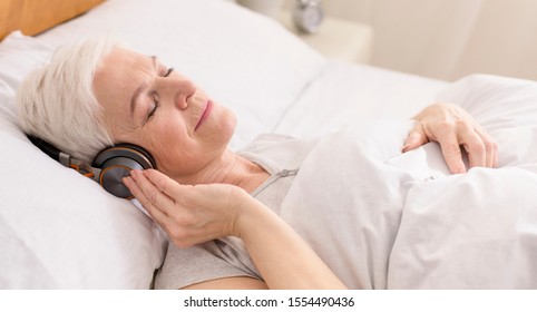 Calm senior woman in headphones enjoying audiobook, resting in bed, panorama
