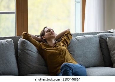 Calm serene sleepy young woman relaxing on comfortable sofa at home, leaning om back, taking deep breath of fresh cool air, enjoying relaxation, leisure time, meditating for stress relief