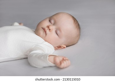 Calm Silent Adorable Baby In White Bodysuit Sleeping On Bed. Cute Few Month Infant Kid Resting On Pale Sheet, Linen. Child Care, Bedtime, Daily Routine Concept. Close Up