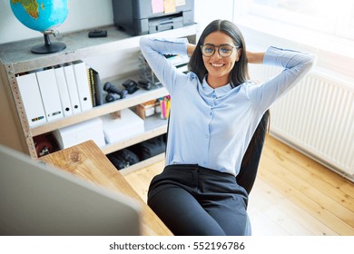 Calm single young woman leaning back in chair at work. Shelf with globe, printer and notebooks behind her.