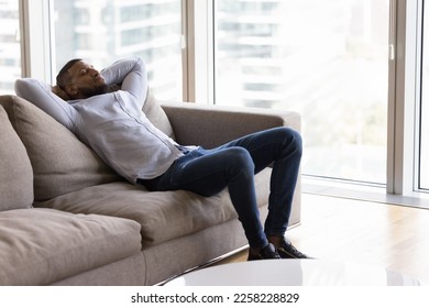 Calm sleepy young African man resting on home sofa, enjoying leisure, silent break. Peaceful Black homeowner guy relaxing on couch in urban apartment with big window, city view in background - Powered by Shutterstock