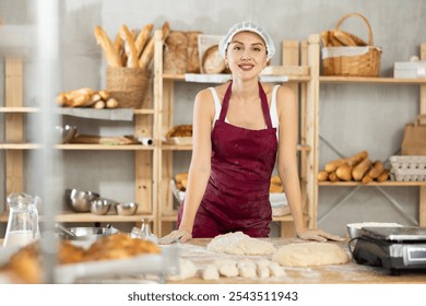 Calm smiling female baker in maroon apron leaning on floured table, enjoying short break while preparing dough in small, well-organized bakery, surrounded by fresh baked goods and tools - Powered by Shutterstock