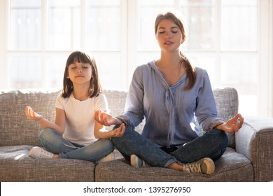 Calm Smiling Mother With Cute Little Daughter Doing Yoga Exercise At Home, Pretty Girl And Attractive Mum With Closed Eyes Sitting In Lotus Pose On Couch, Relaxing Together, Family Exercise