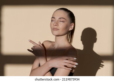 Calm Thoughtful. Young Beautiful Slim Caucasian Woman In Black Swimsuit Isolated Over Brown Studio Background. The Concept Of Beauty, Body And Skin Care, Healthy Lifestyle, Sun Shadows