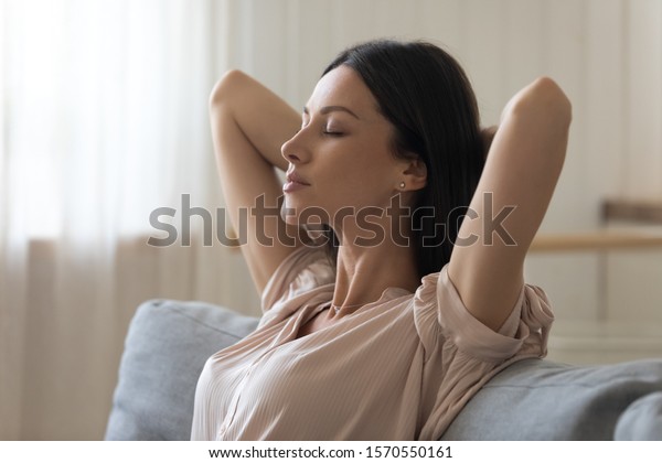 Calm tired young woman attractive face lounge\
leaning on sofa napping with eyes closed at home, serene lady hold\
hands behind head relaxing breathing fresh air sit on comfortable\
couch in living room