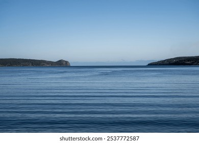 A calm tranquil ocean with ripples between two landmassses. The calm sea is deep blue. The sky is blue and the sun is setting on the horizon. The land is a rocky coastline enclosing the peaceful cove. - Powered by Shutterstock