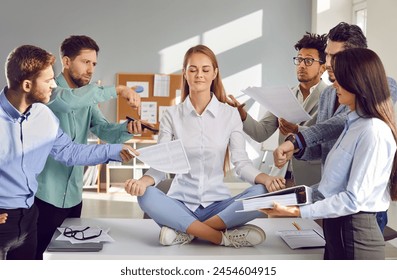 Calm, tranquil woman sitting on office table in lotus yoga pose, mediating, chilling out, and ignoring team of diverse colleagues who are demanding to do lots of work as soon as possible - Powered by Shutterstock