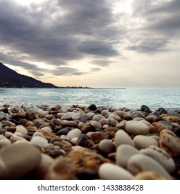 Calm Water At Coast. Ripple Of Water Near Shoreline. Silhouette Of City On Skyline. Stone Seashore Before Storm. Rocky Beach Near The Bay.