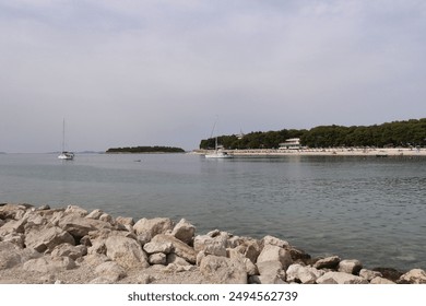 Calm waters of the bay stretch out under a cloudy sky, with two sailboats gently anchored near a rocky shoreline and a distant forested coastline. - Powered by Shutterstock