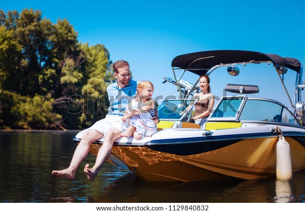 Calm weekend. Pleasant young family\
relaxing on a motorboat while petite little girl and her father\
sitting on the bow of the boat and the mother\
sailing