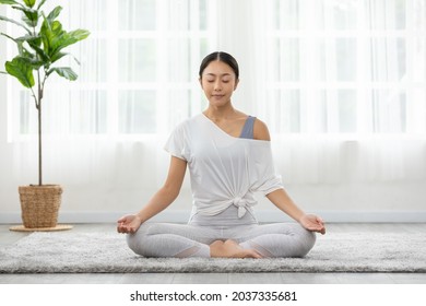 Calm of wellness Asian young woman sit on carpet breathing with yoga lotus pose,Yoga meditation of young healthy woman relax and comfortable at white cozy home,Yoga Exercise for Wellness Concept - Shutterstock ID 2037335681