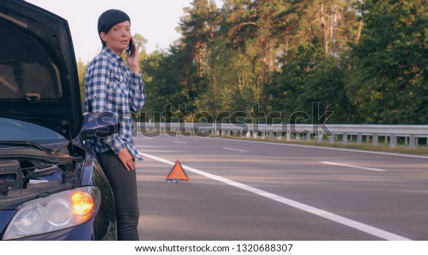 Calm woman calling roadside assistance for help\
after incidents. Young casual girl using smartphone near broken\
down car turn on hazard\
lights.