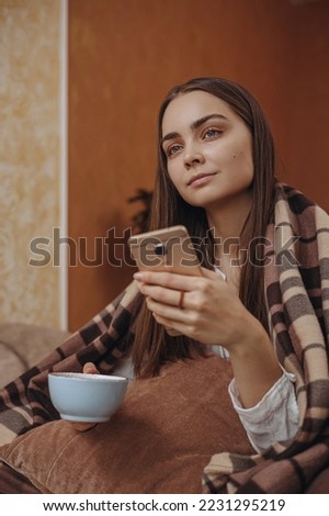 Calm woman drinking tea and using smartphone while relaxing in cozy plaid at home and looking away dreamily 