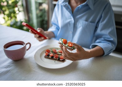 Calm woman leisurely eating rye crisp bread toast sandwich with cream cheese, cherry, arugula, basil while using mobile phone. Soft focus on female hands with food, smartphone, checking social network - Powered by Shutterstock